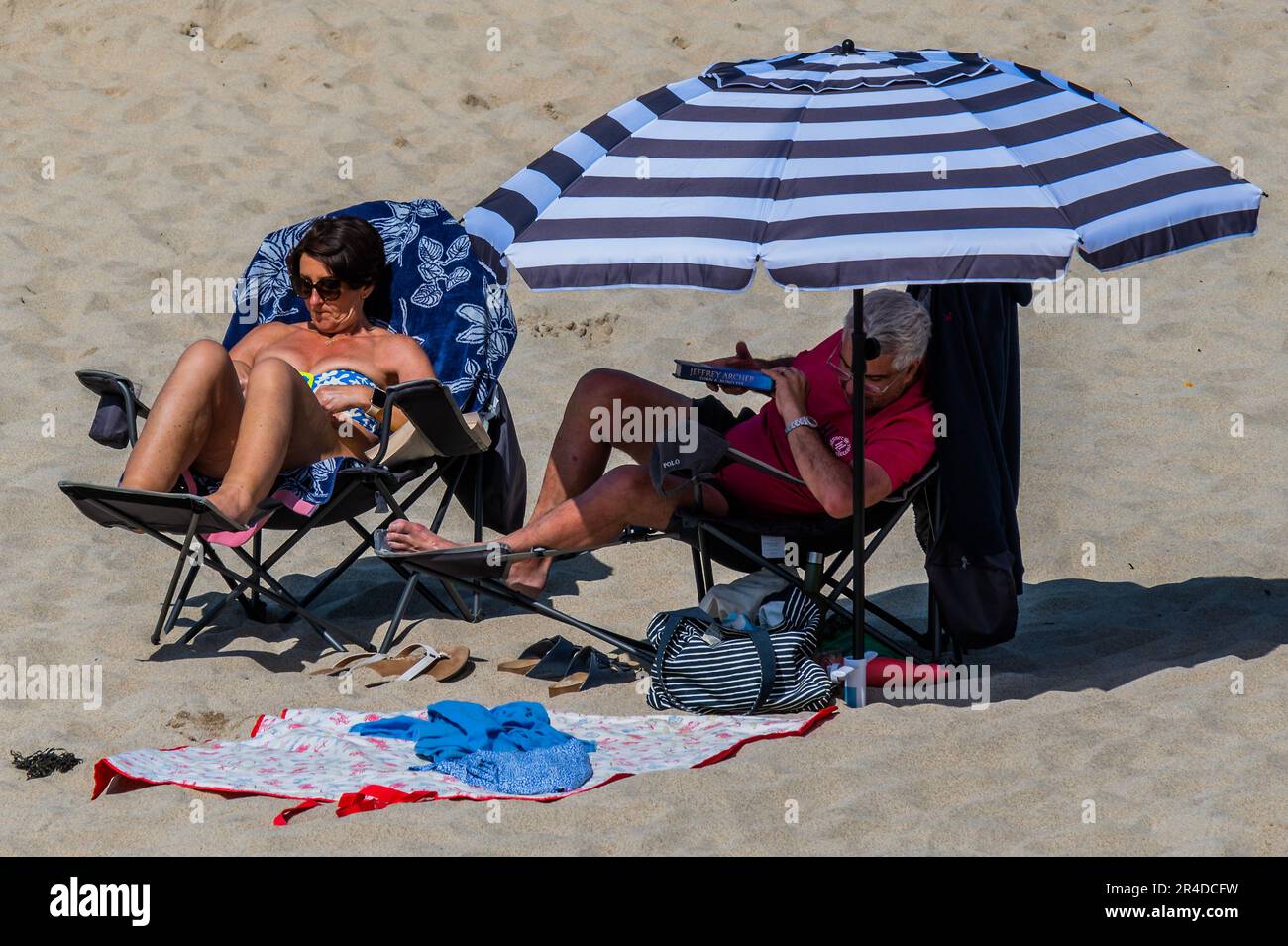 St Ives, UK. 27th May, 2023. A man puts down the latest Jeffry Archer book under a huge sunshade, while his friend reads in the sun next to him - People flock to the beach to sunbathe as well as take part in watersports and boat trips - Sunny weather for the bank holiday weekend in St Ives. Credit: Guy Bell/Alamy Live News Stock Photo