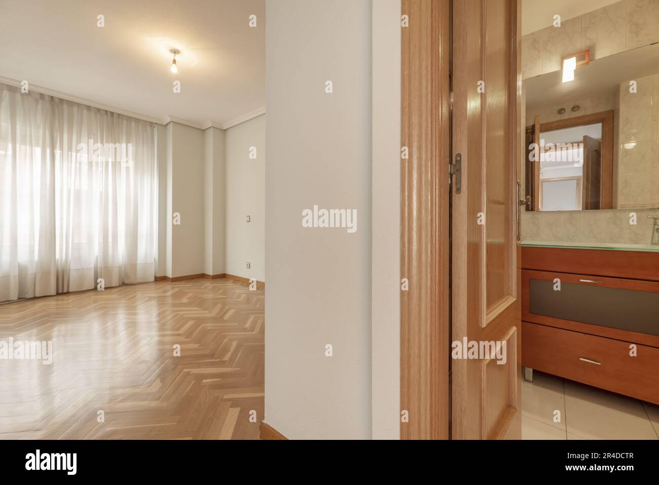 An empty room with a varnished oak parquet floor with a large window with white curtains and an en-suite bathroom Stock Photo