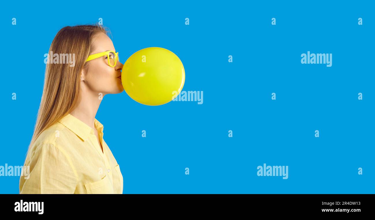 Happy young woman blowing up yellow balloon on blue copy space banner background Stock Photo