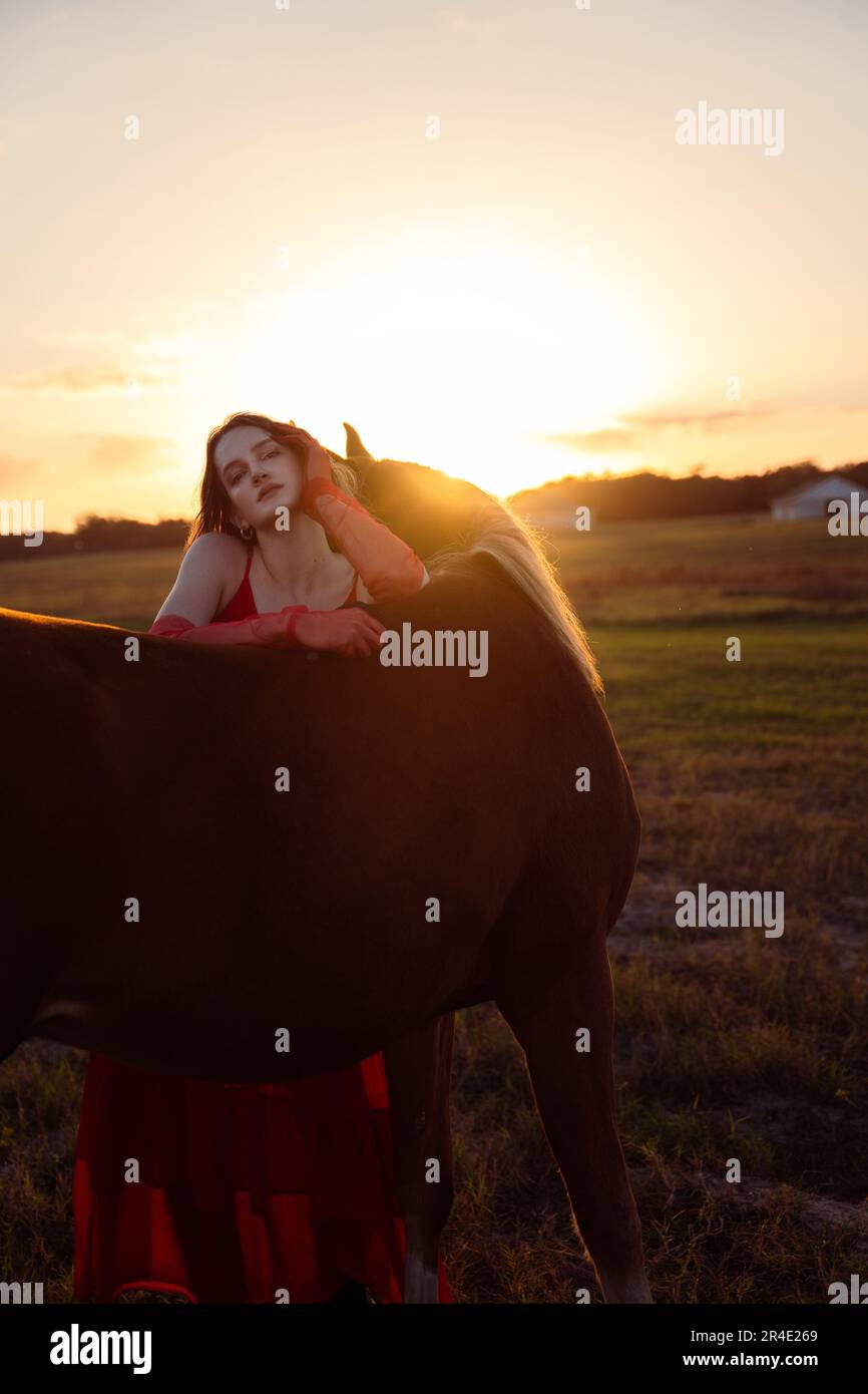 A beautiful woman in a red dress posing in a meadow with a horse at ...