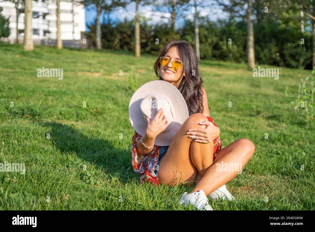 Joyful woman with sunglasses looking at camera and smiling while sitting outdoors on a green grass in a park. Stock Photo