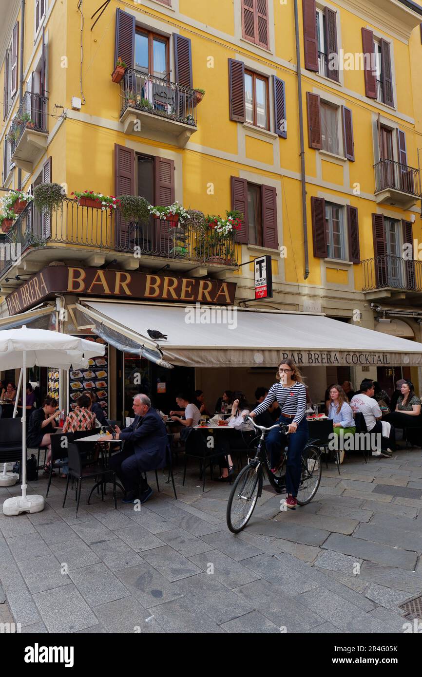 Lady rides a cycle beside a yellow building with restaurant in the chic Brera district of Milan, Lombardy, Italy Stock Photo