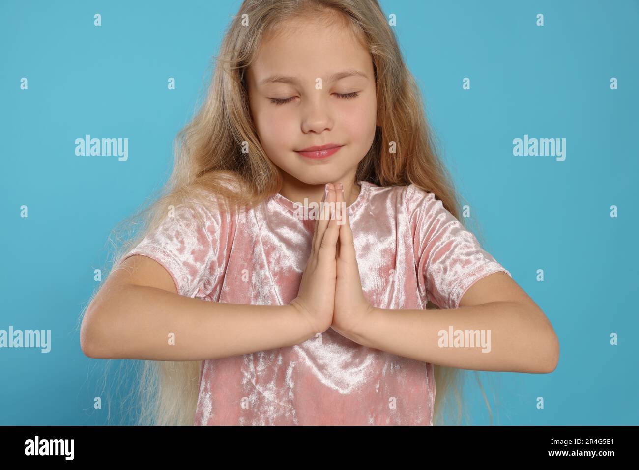 Beautiful girl with hands clasped together and closed eyes praying on light blue background Stock Photo