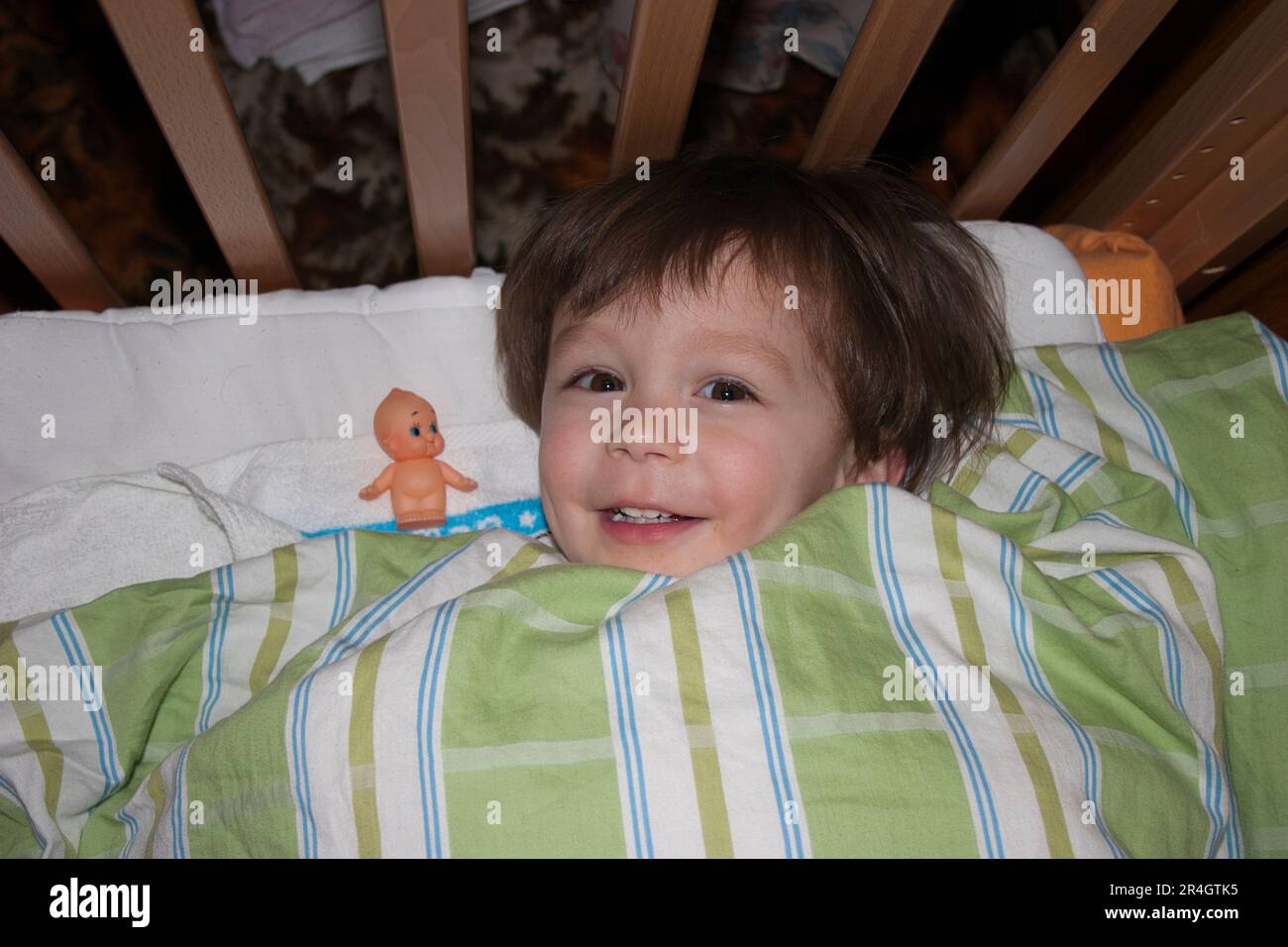 One year old Caucasian child, boy, laying in cot with duvet over him, with his 'friend,' a kewpie doll next to him. Eye-contact. Smiling. Stock Photo