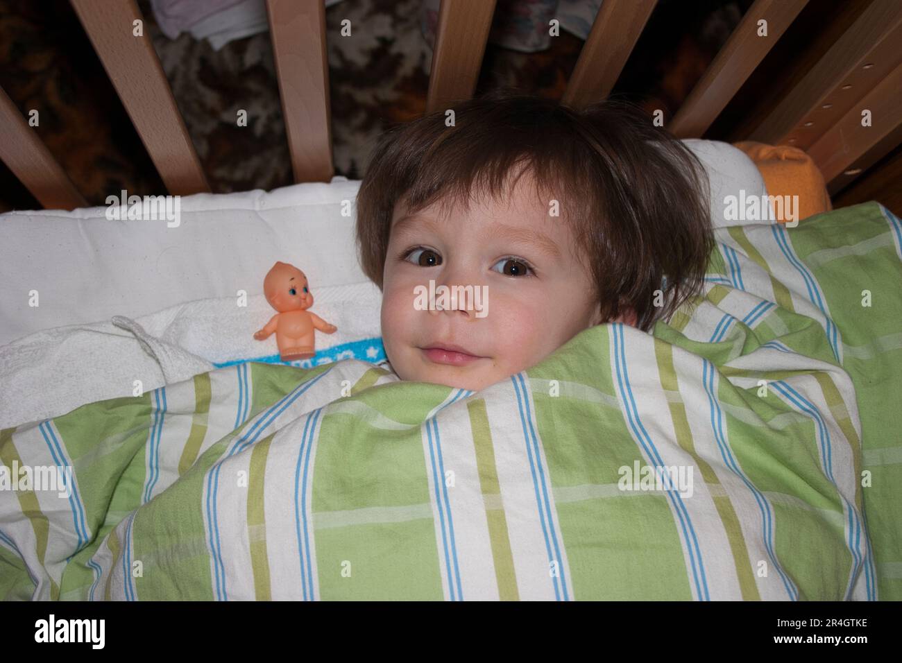 One year old Caucasian child, boy, laying in cot with duvet over him, with his 'friend,' a kewpie doll next to him. Eye-contact. Stock Photo