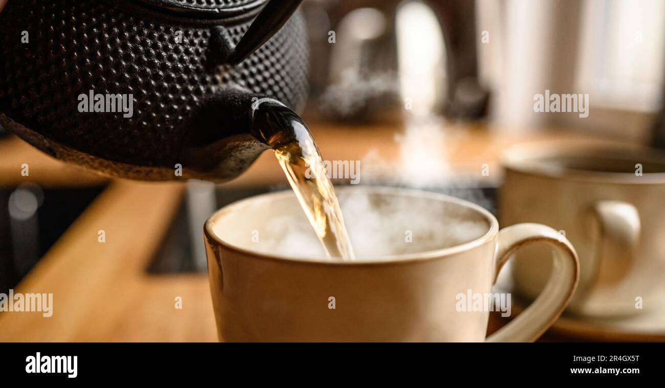 Pouring hot black tea from metal teapot at the kitchen. Steam comes out of the cup. T Stock