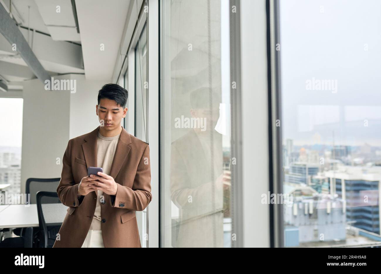 Young Asian business man using smartphone standing in office at window. Stock Photo