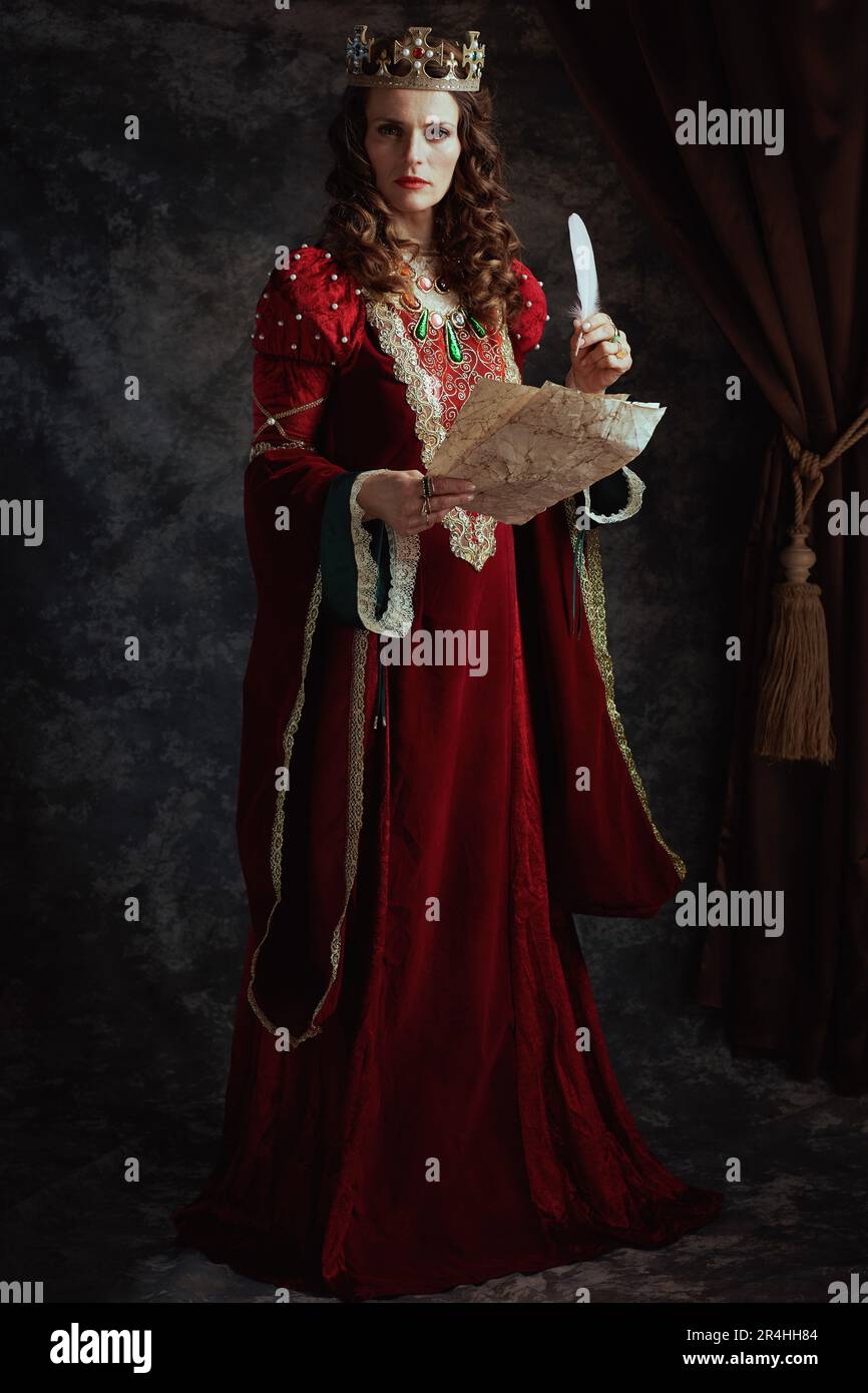 Full length portrait of medieval queen in red dress with parchment and crown on dark gray background. Stock Photo
