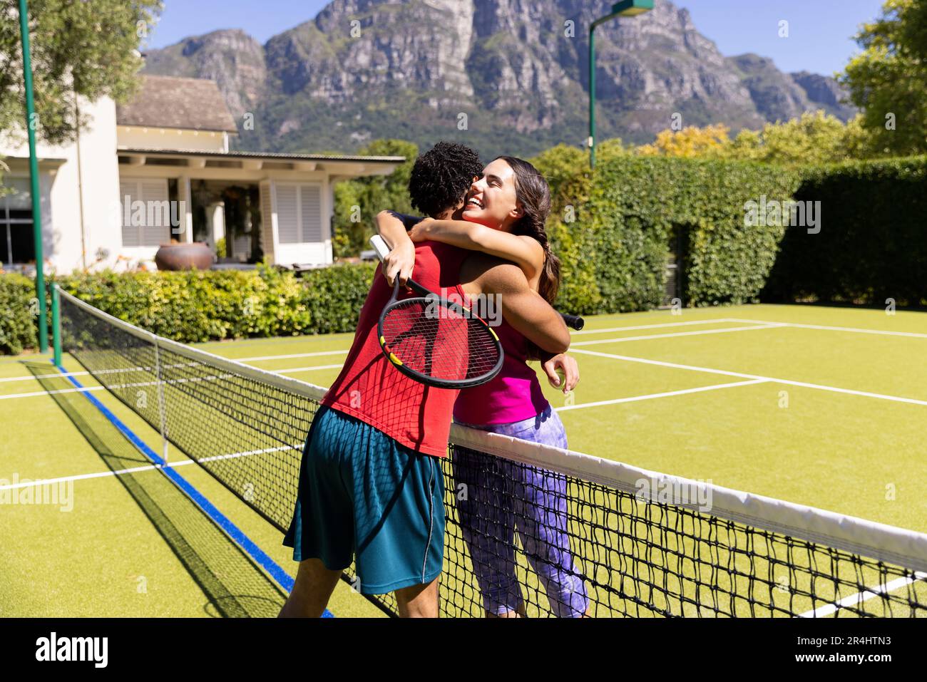 Biracial happy young couple with tennis rackets embracing while standing in court after game Stock Photo