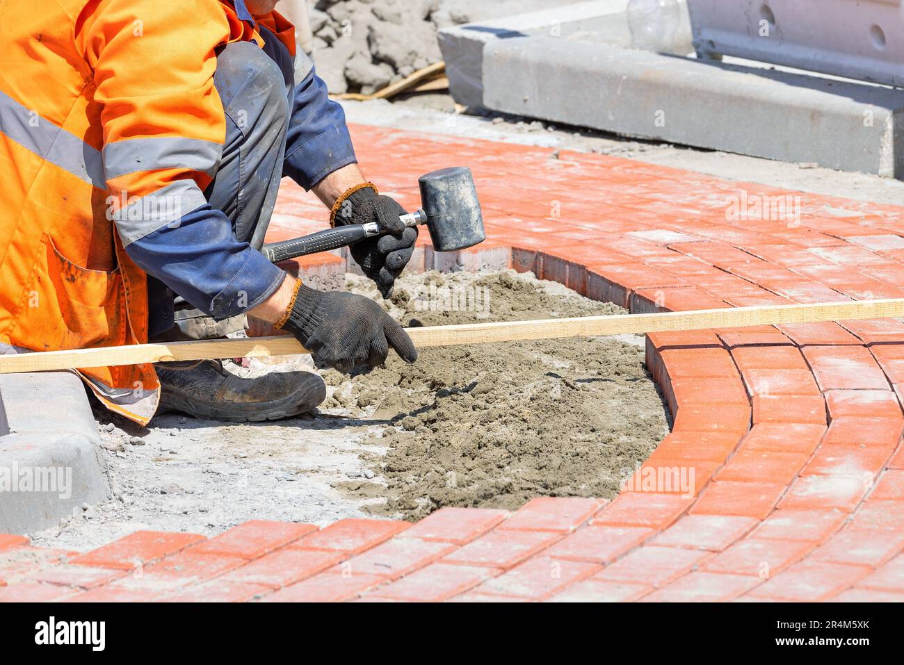 A bricklayer in work gloves and overalls carefully measures the size with a wooden plank and lays orange paving slabs on a footpath on a sunny day. Stock Photo