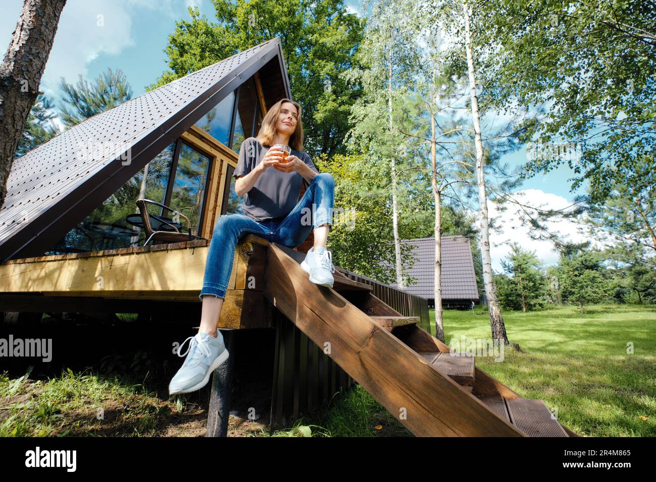 Happy woman sits on terrace of log cabin with a cup of tea and enjoys ...