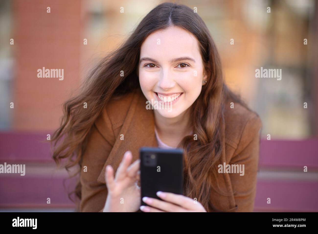 Happy woman holds smart phone looking at you in winter on a bench in the street Stock Photo