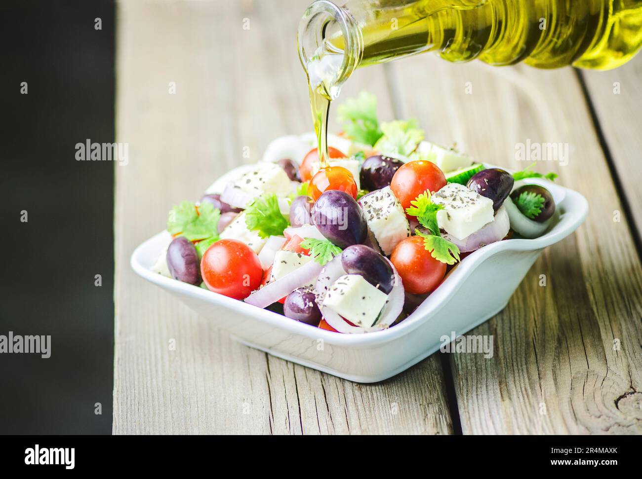 Fresh olive oil pouring into bowl of Greek Salad with tomatoes, feta cheese, onion, parsley, black olives and  oregano on wooden background.. Stock Photo