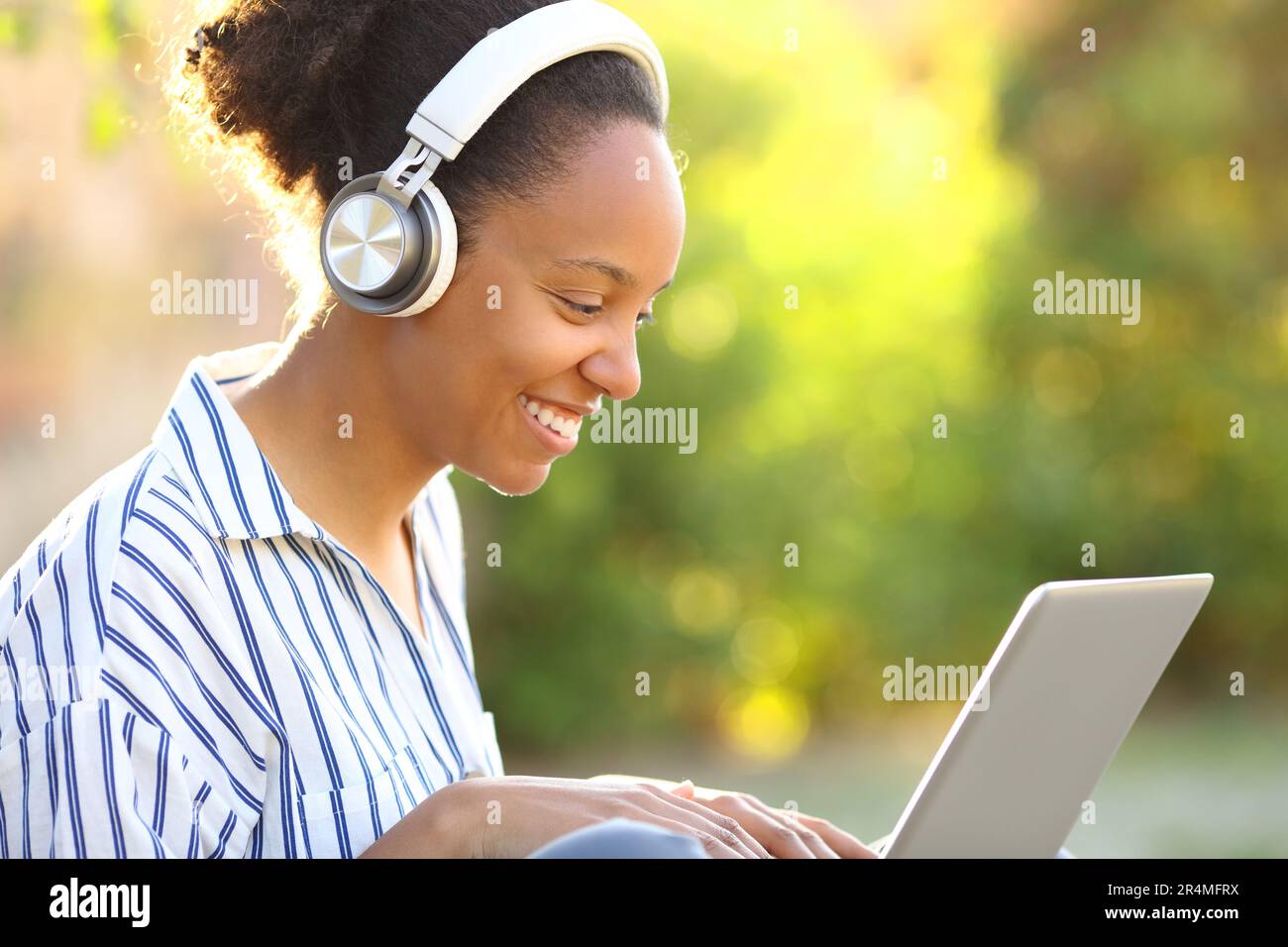 Happy black woman watching media on laptop with headphone in a green