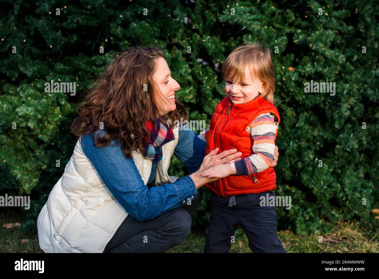 A laughing mother and small child sit together in front of lit-up tree Stock Photo