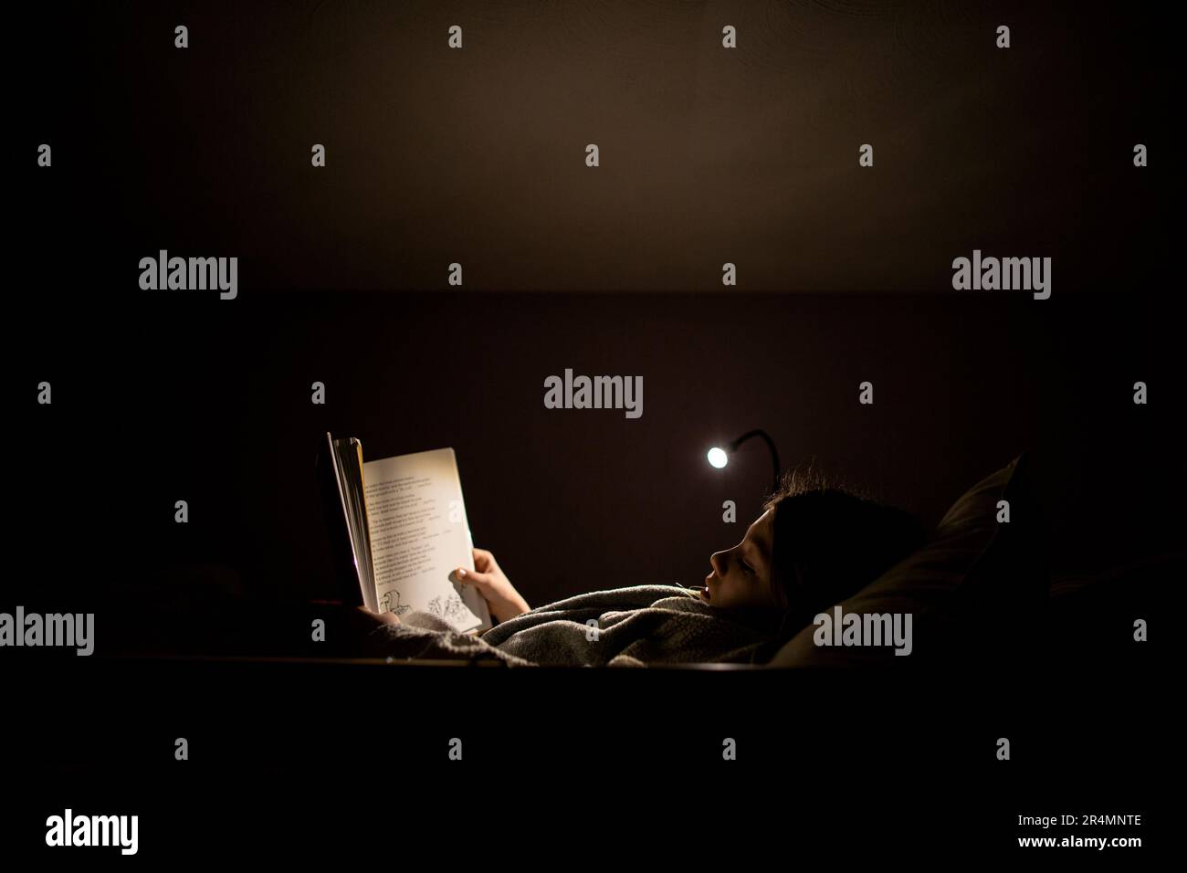 Backlit image of young girl reading by lamp light in bed at nighttime