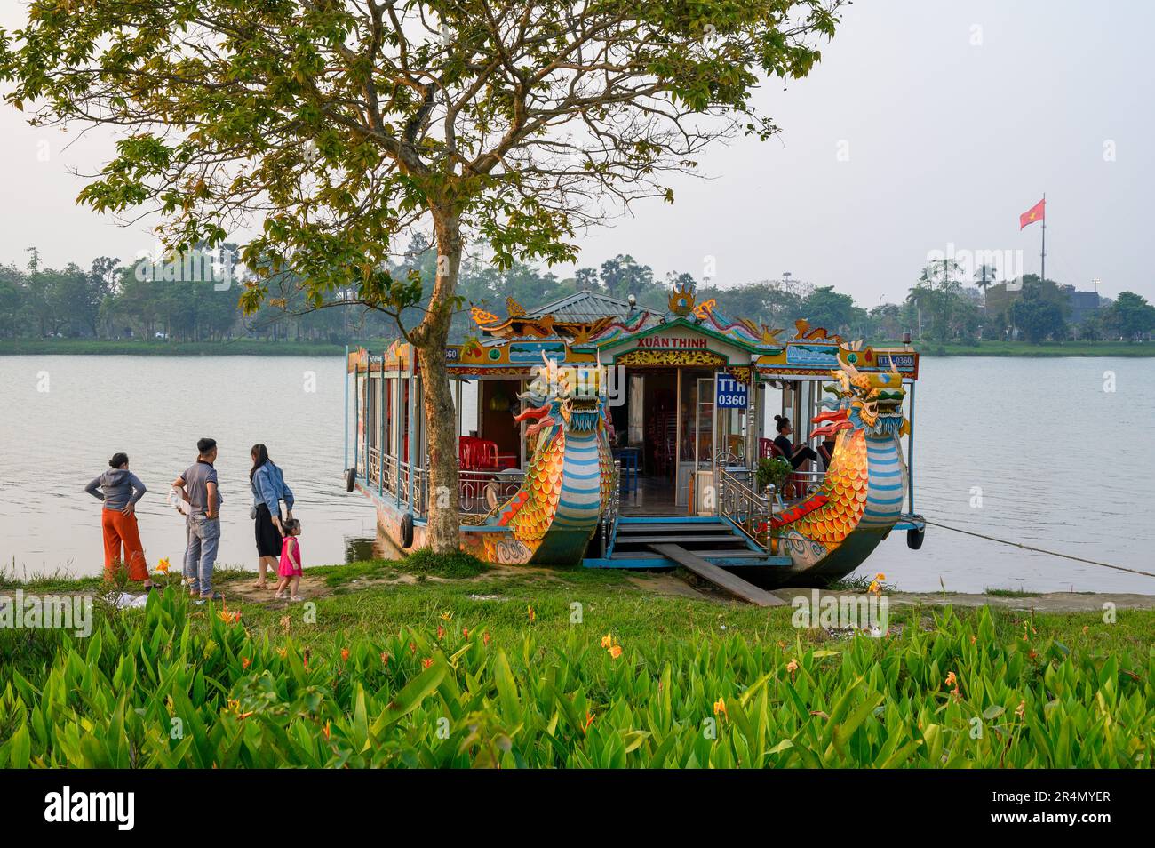 A dragon boat moored on the Perfume River and a local family going about everyday life on the riverbank in Hue, Vietnam. Stock Photo