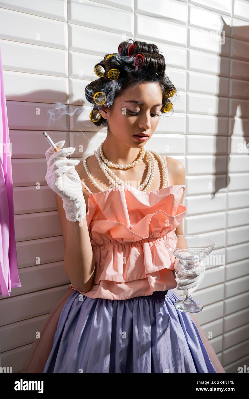 asian young woman with hair curlers standing in pink ruffled top, pearl necklace and gloves while holding cigarette and glass near wet laundry and  wh Stock Photo