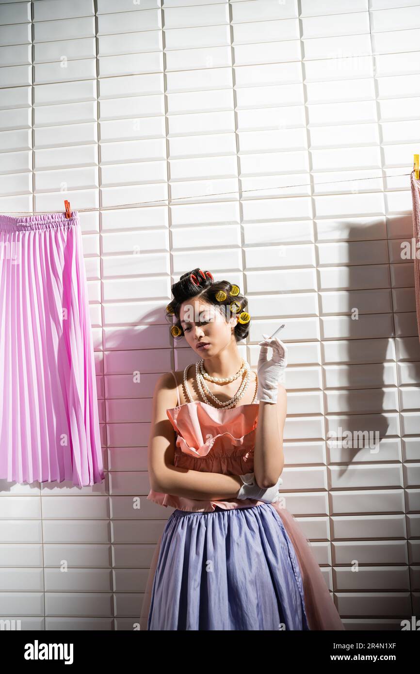 asian young woman with hair curlers standing in pink ruffled top, pearl necklace and gloves while holding cigarette near wet laundry hanging near whit Stock Photo