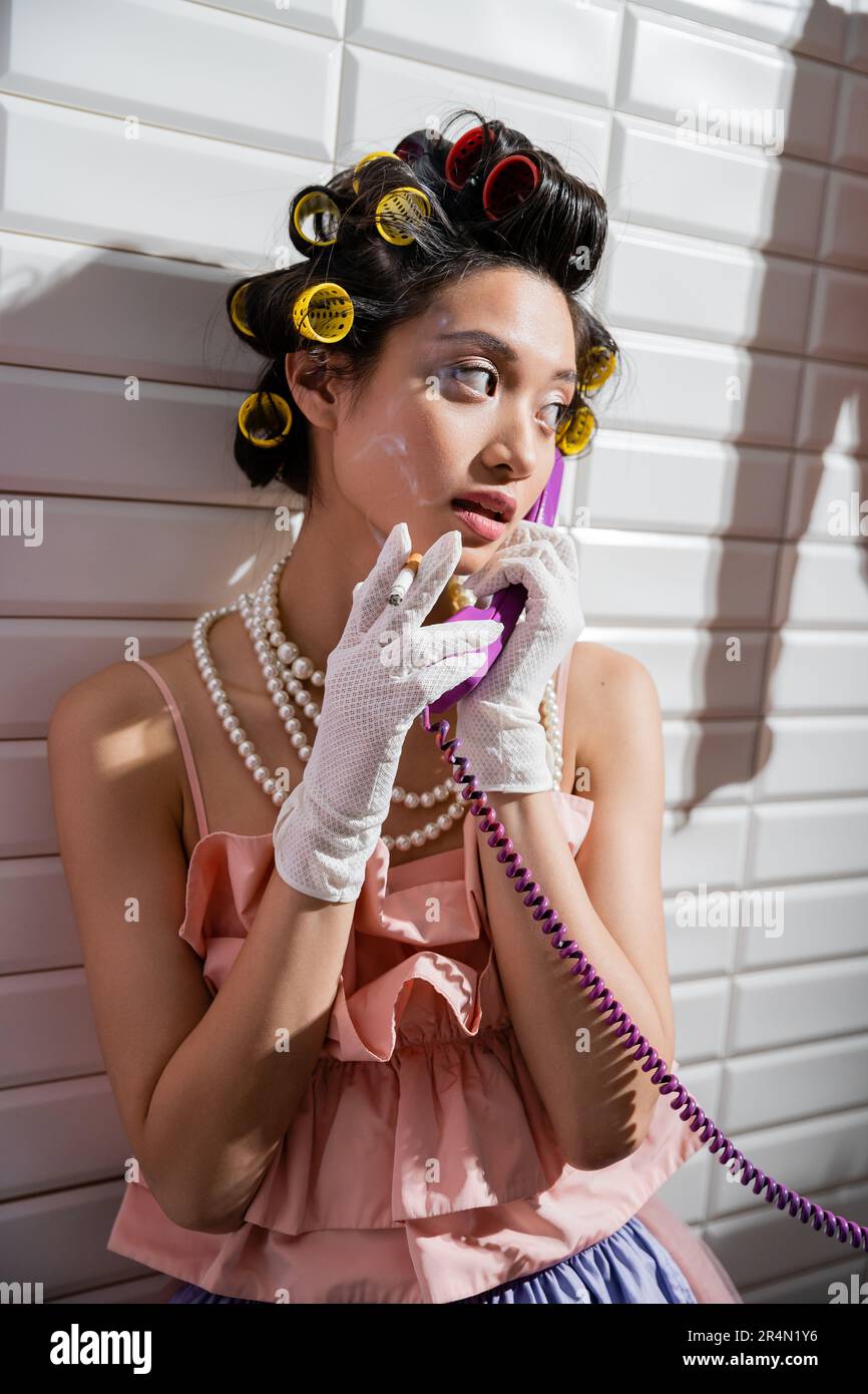 brunette and asian young woman with hair curlers standing in pink ruffled top, pearl necklace and white gloves, smoking cigarette and talking on retro Stock Photo