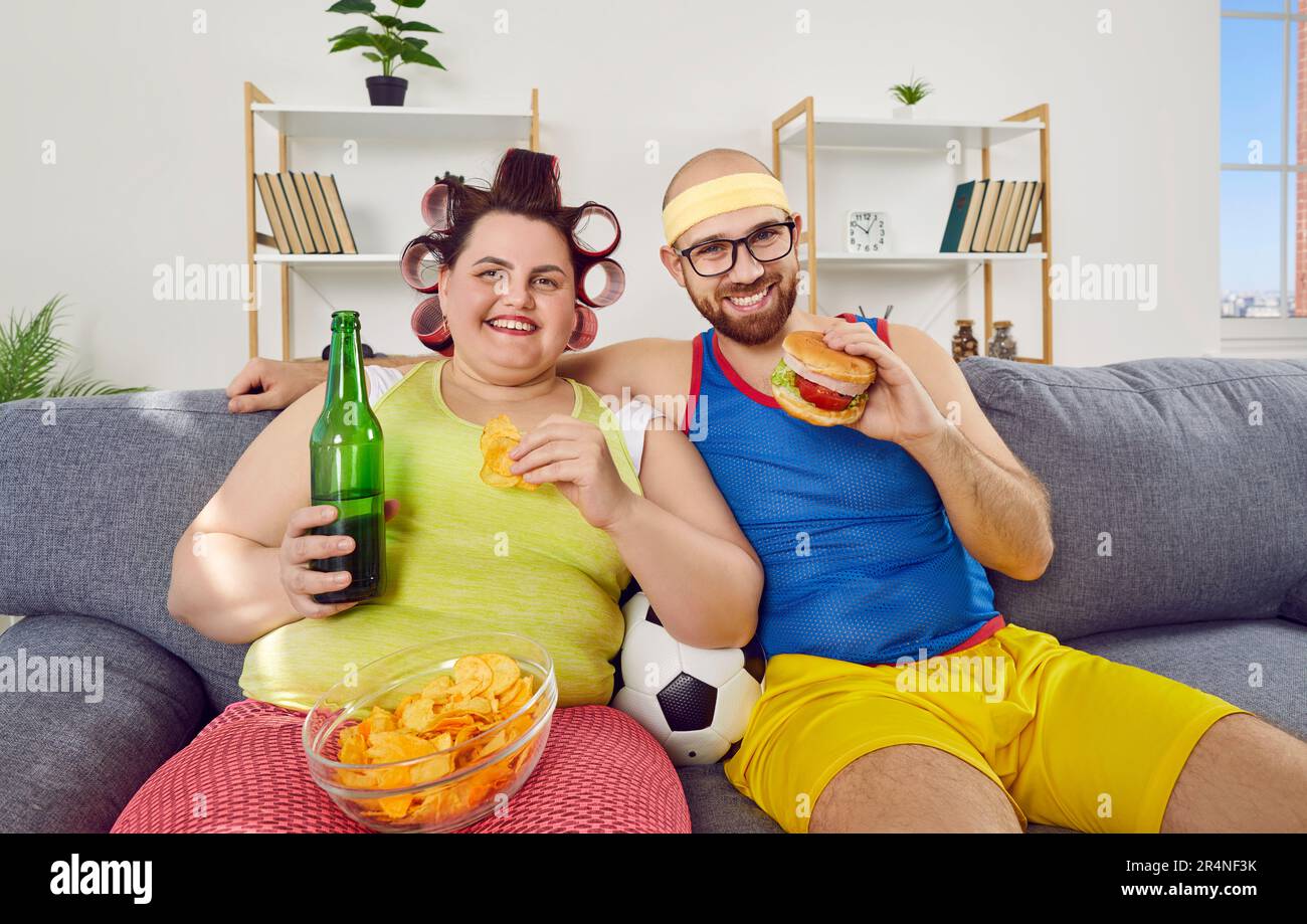 Funny couple watching football sport game sitting on sofa at home with beer and fast food. Stock Photo