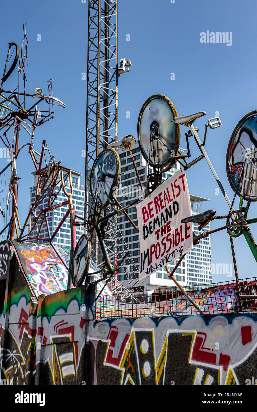 'Be realist, demand the impossible' sign among scrapped bikes at the Suvilahti DIY skatepark. Kalasatama towers in the background. Stock Photo