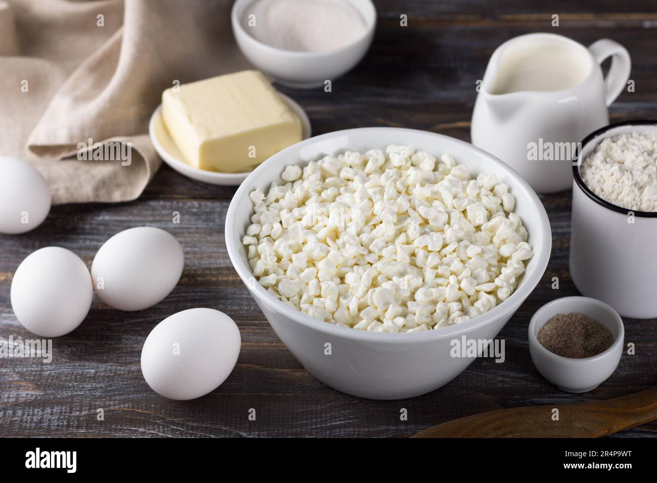 Bowl with cottage cheese, eggs, flour, butter, milk, sugar, vanilla sugar on a wooden background. Ingredients for home baking Stock Photo