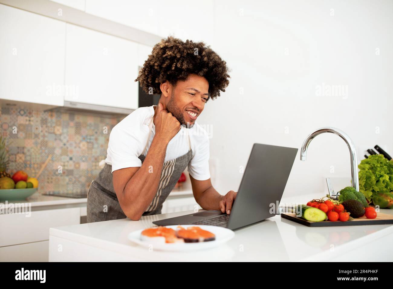 Chef Preparing Dinner Using Laptop For Recipes Search At Kitchen Stock Photo