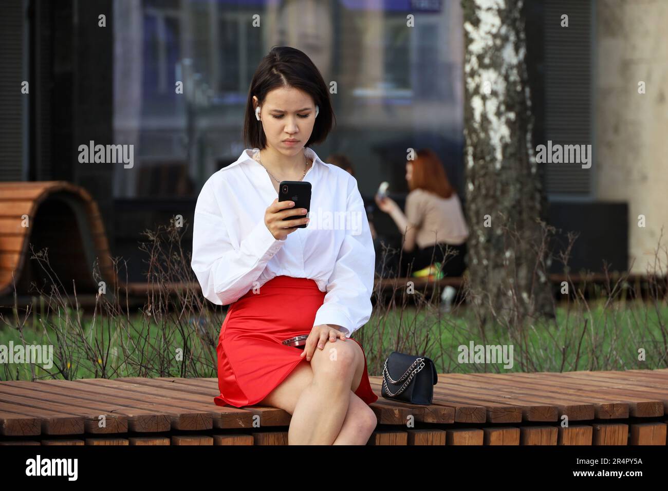 Girl in a white blouse and a red skirt using smartphone sitting on wooden bench on city street Stock Photo