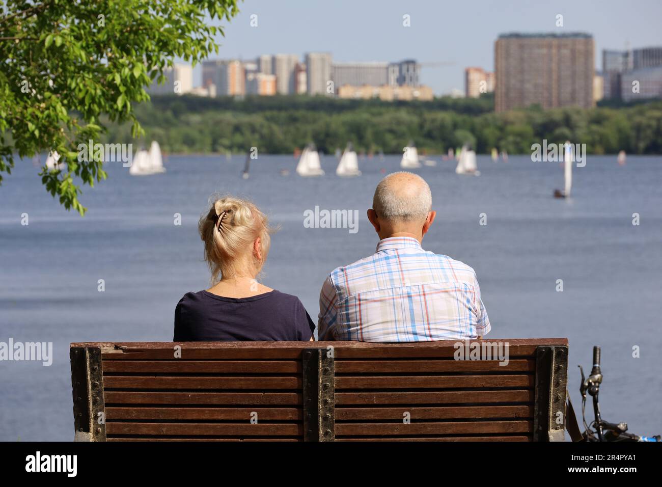 Old woman and man sitting on a wooden bench on river and city background, rear view. Couple of retirees on a beach, relax and enjoying life in summer Stock Photo