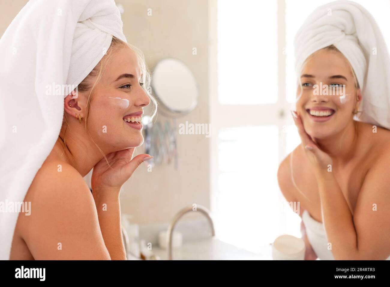 Plus size caucasian woman with towel wrapped on head applying cream on face and looking in mirror Stock Photo