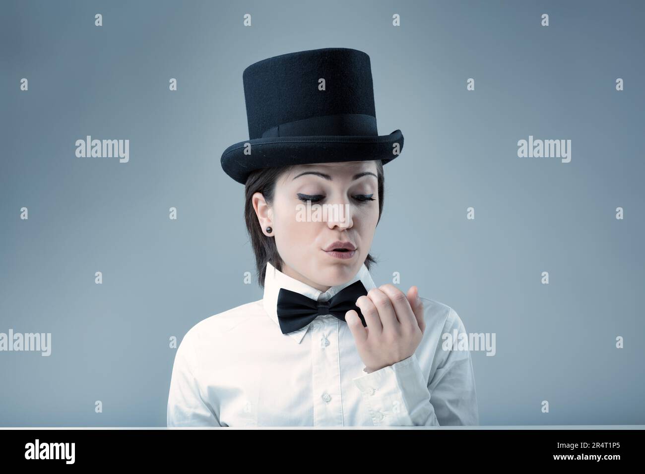 Woman in top hat, air of superiority, blowing on her nails, demands nothing less than her standards. White shirt, black bow tie, neutral background Stock Photo