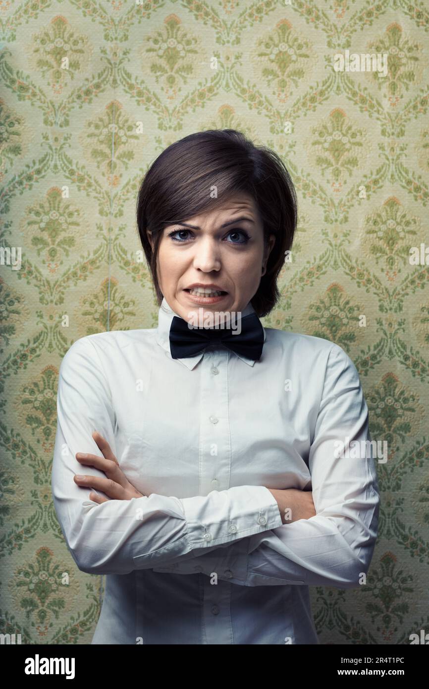 Woman with crossed arms wearing a white shirt and black bow tie in front of a vintage wallpaper background. Her doubtful and annoyed expression sugges Stock Photo