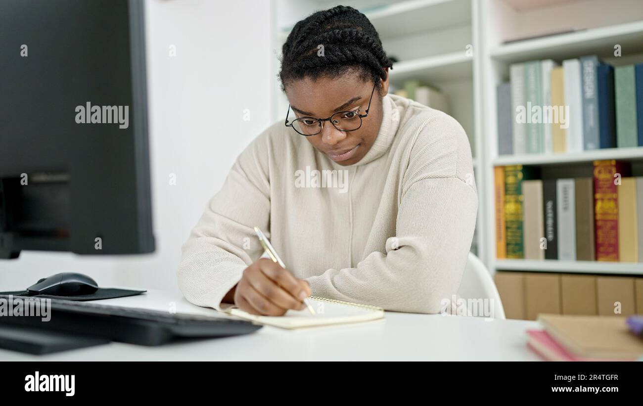 African american woman student using computer writing notes at library ...