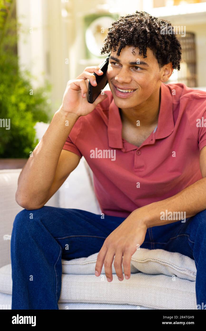 Biracial smiling young man talking over smartphone while sitting on sofa in living room Stock Photo