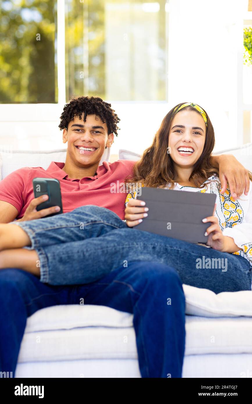 Portrait of happy biracial young couple using smartphone and digital tablet while relaxing on sofa Stock Photo