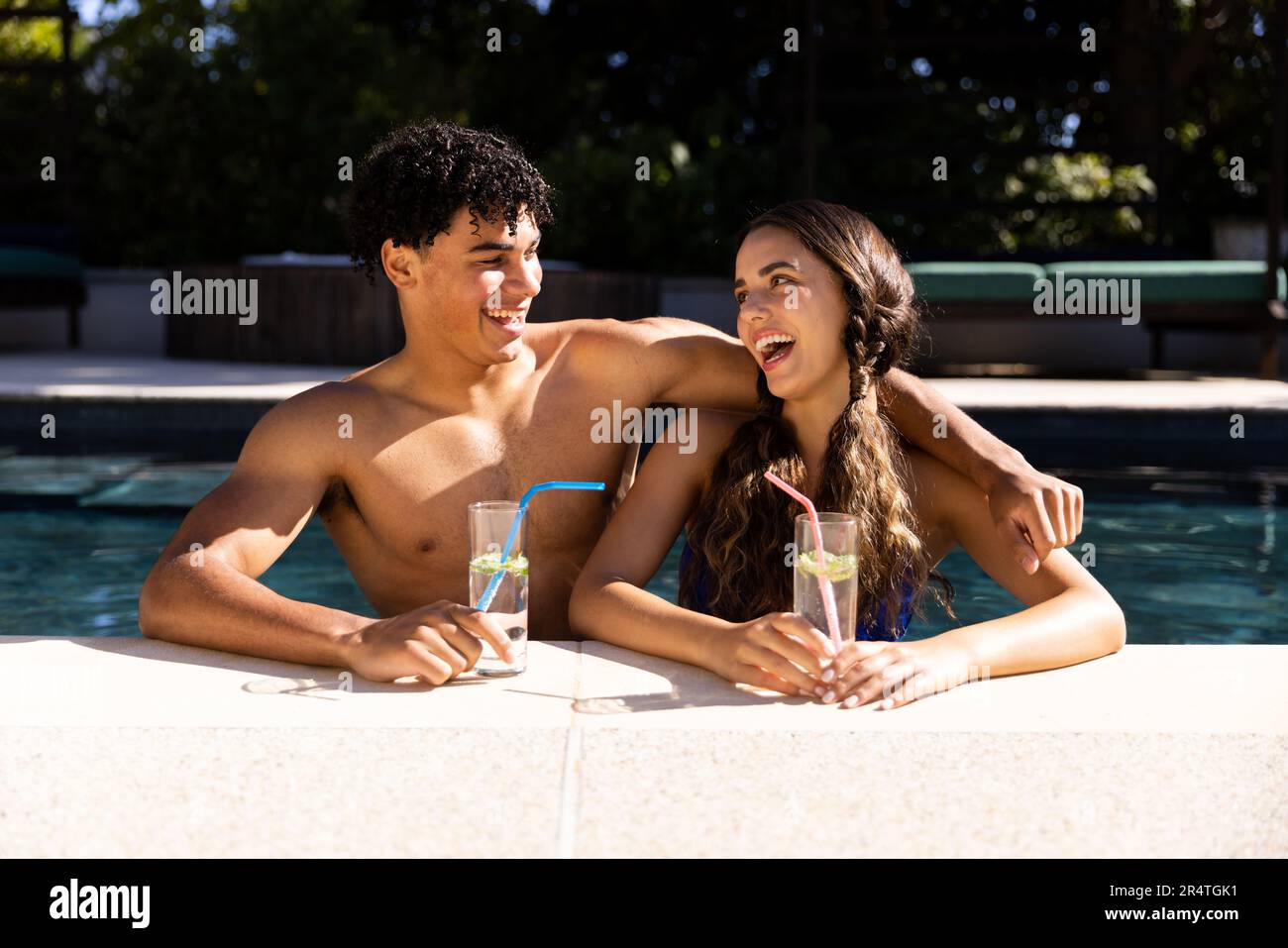 Happy biracial young couple holding drinks and talking in swimming pool during sunny day Stock Photo