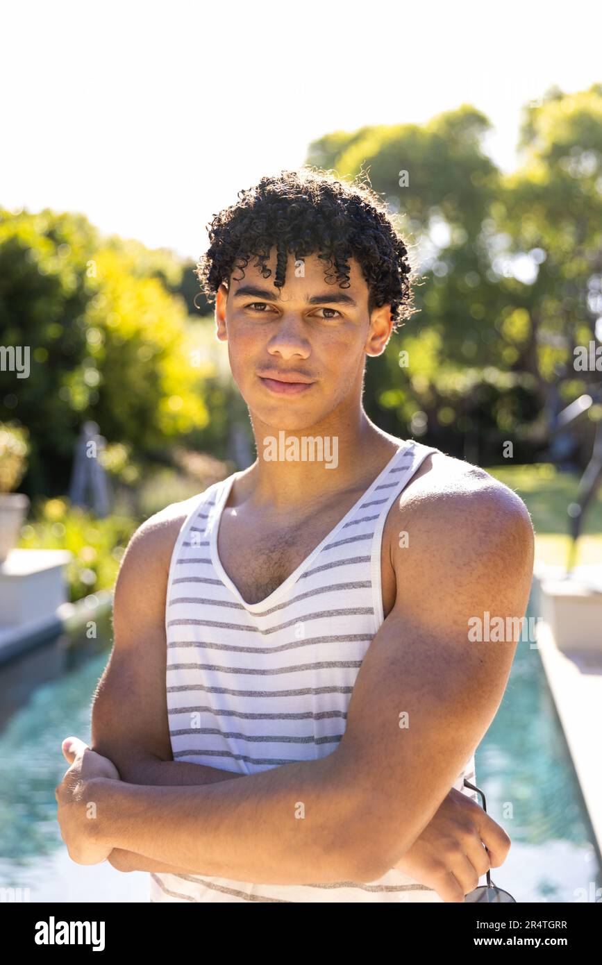 Portrait of biracial young man holding sunglasses and standing at poolside against clear sky Stock Photo