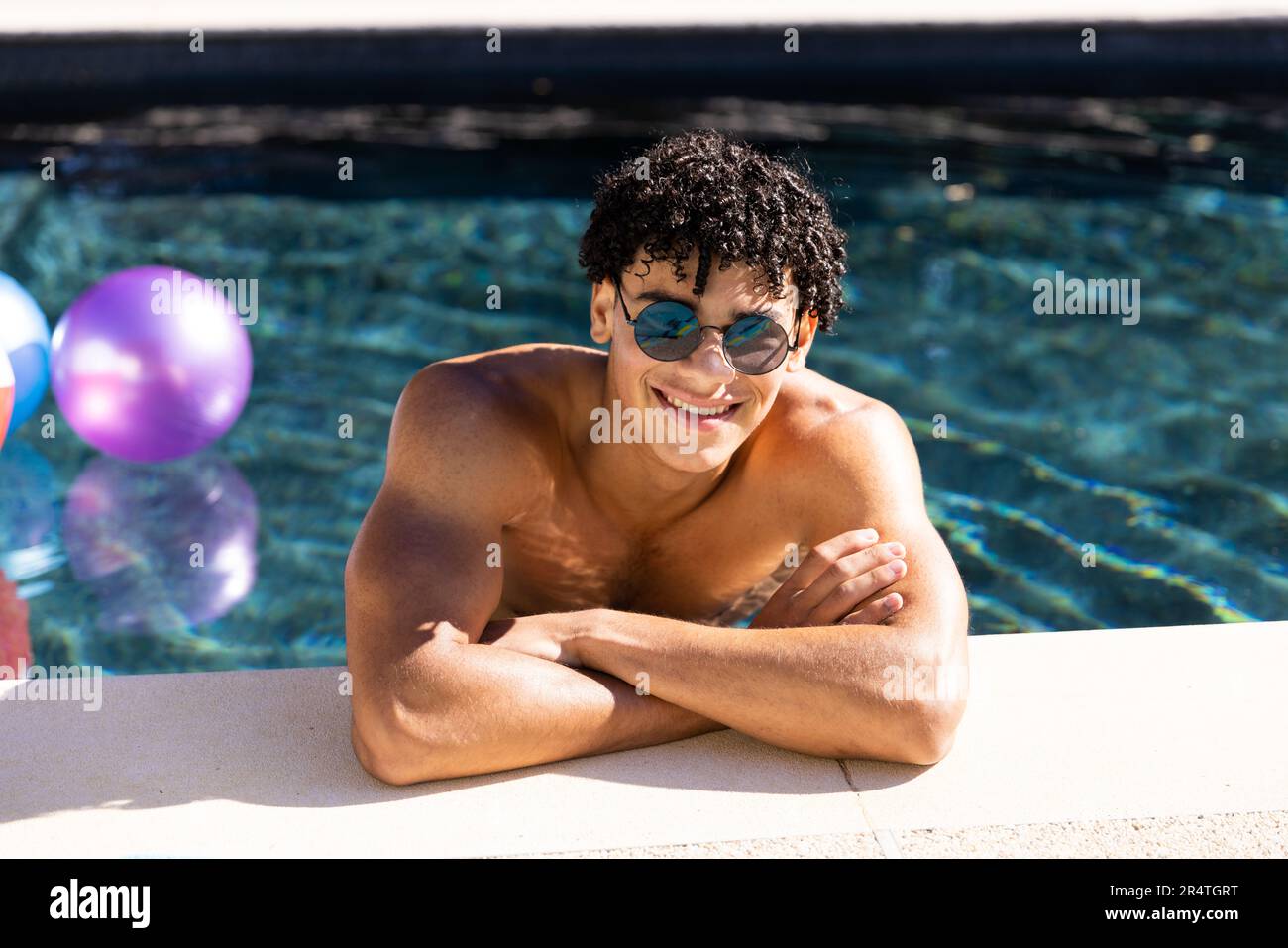 Portrait of biracial shirtless young man in sunglasses smiling and leaning at poolside Stock Photo