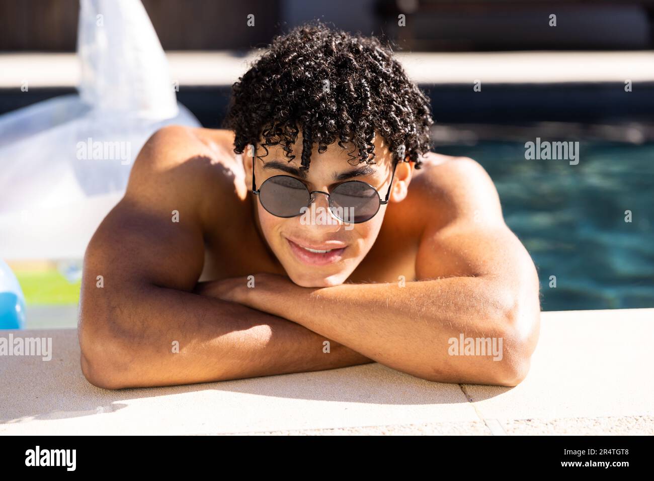 Portrait of biracial shirtless young man in sunglasses with arms crossed leaning at poolside Stock Photo