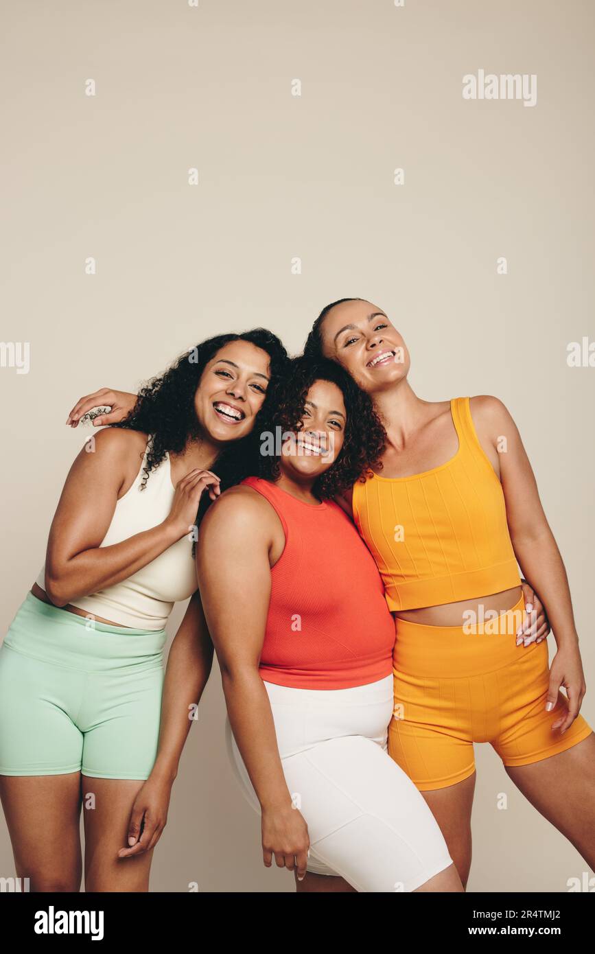 Group of sporty women smiling at the camera, standing in a studio in fitness clothing. Three young female friends embracing a lifestyle of sport, exer Stock Photo