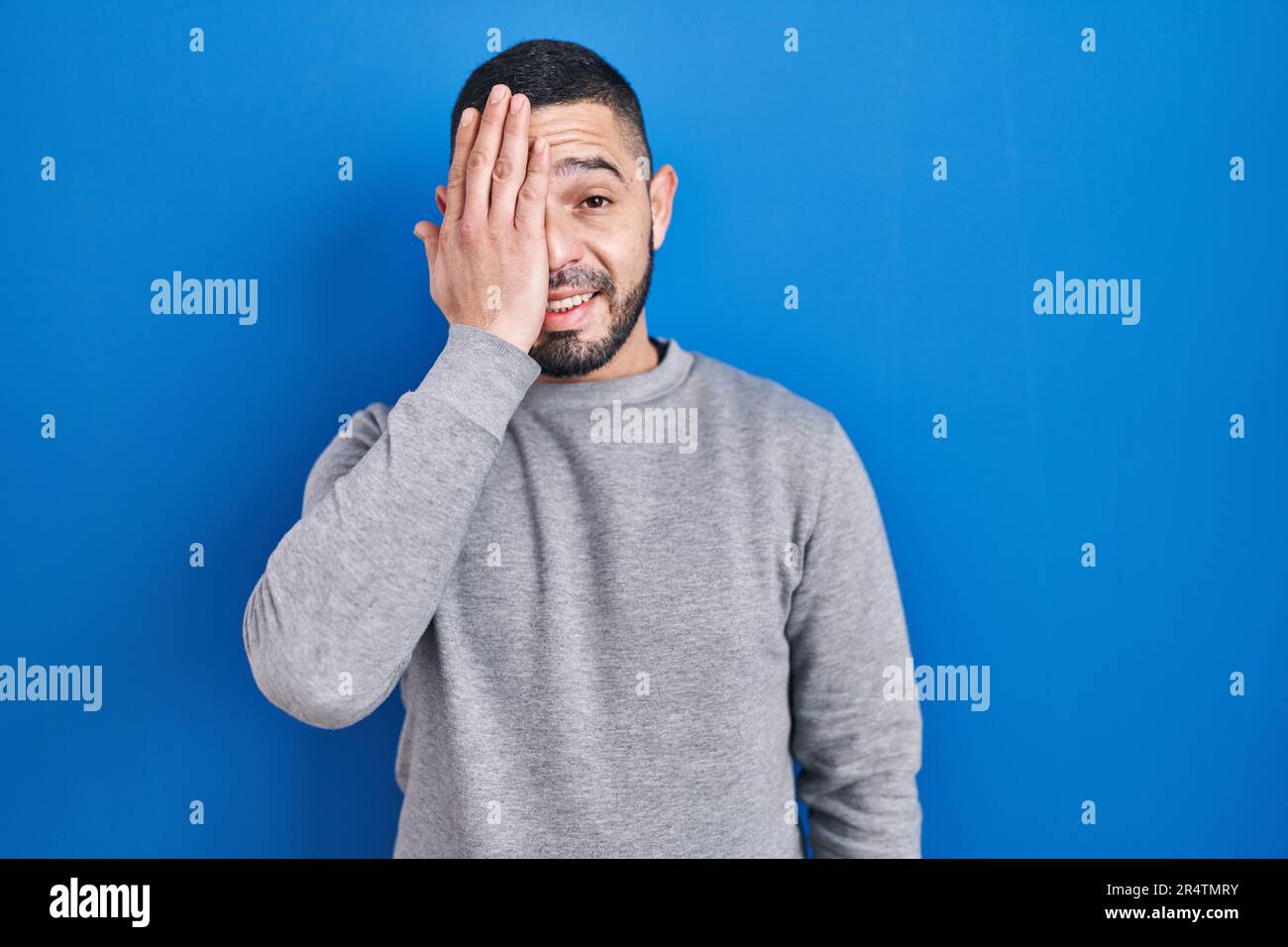 Hispanic man standing over blue background covering one eye with hand ...
