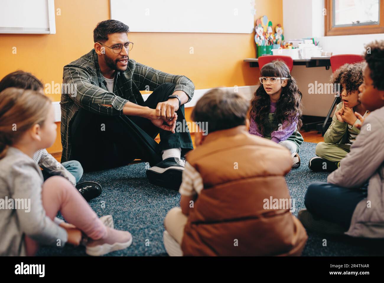 Teacher sitting on the floor with his students in an elementary school classroom, he is talking to them as they sit in a circle. Man teaching a group Stock Photo