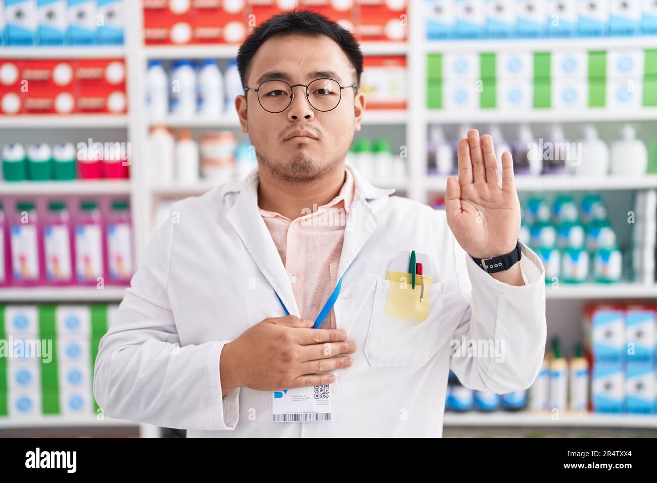 Chinese young man working at pharmacy drugstore swearing with hand on chest and open palm, making a loyalty promise oath Stock Photo