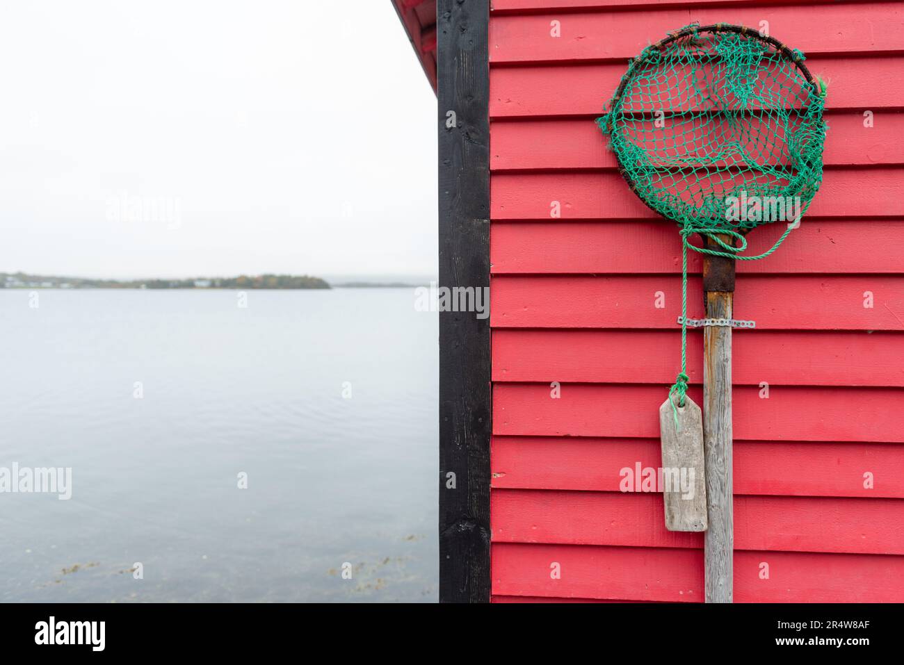 A vintage fishing dip net hanging on a vibrant red wooden horizontal clapboard wall. The scoop is rusty and the cloth mesh rope net is torn and worn Stock Photo