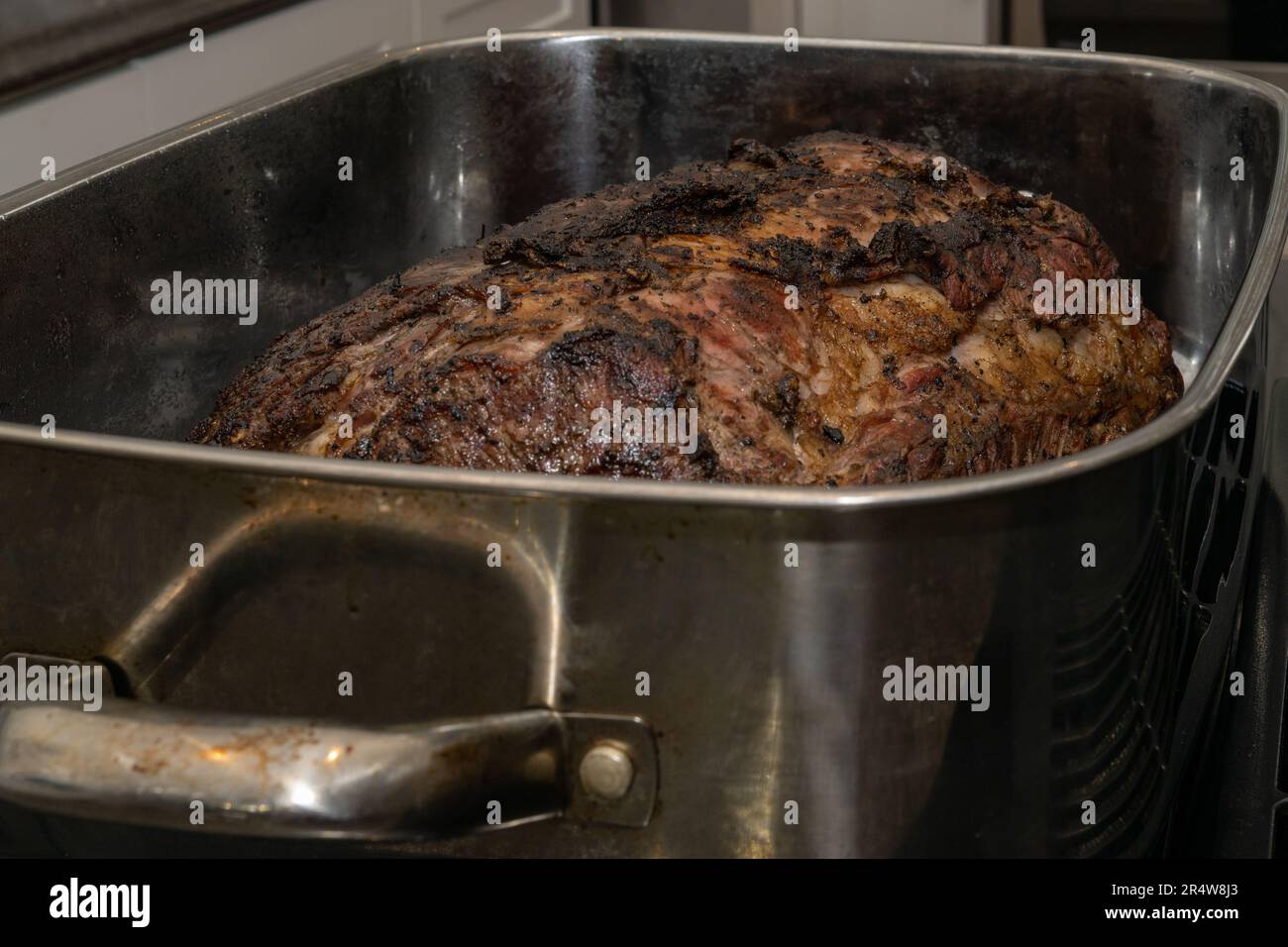 A large well-done Angus beef prime rib roast cooked in a roaster pan with onions and garlic. The shiny metal pan is rectangular in shape with handles. Stock Photo