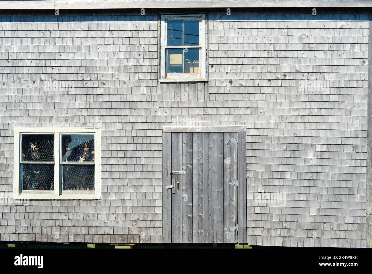 The exterior of a worn grey weathered wooden wall of a shed or barn. There are two windows with four panes of glass and a plank door with a latch. Stock Photo