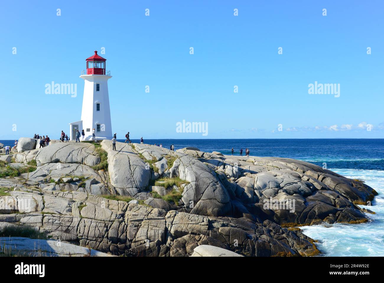 Scenic Peggy's Cove, with a tall white lighthouse with a red tower on top. The concrete structure has three windows. There are people walking below. Stock Photo