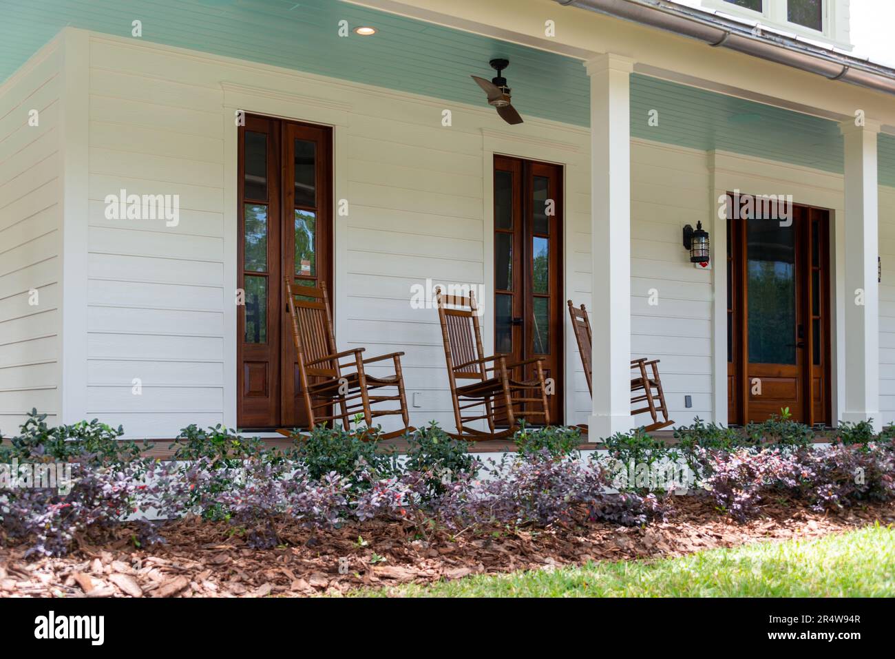 Three vintage brown wooden rocking chairs on the veranda of an old white colored country house with a wrap around porch. There's a flower bed. Stock Photo