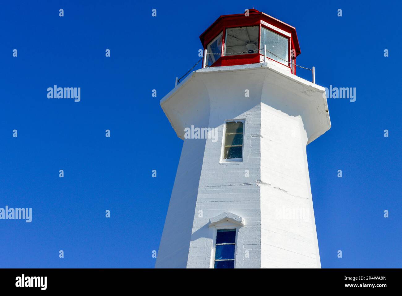 A vintage octagon shaped white concrete lighthouse with two small glass windows, a red metal watch tower with a green light, and a metal rail. Stock Photo